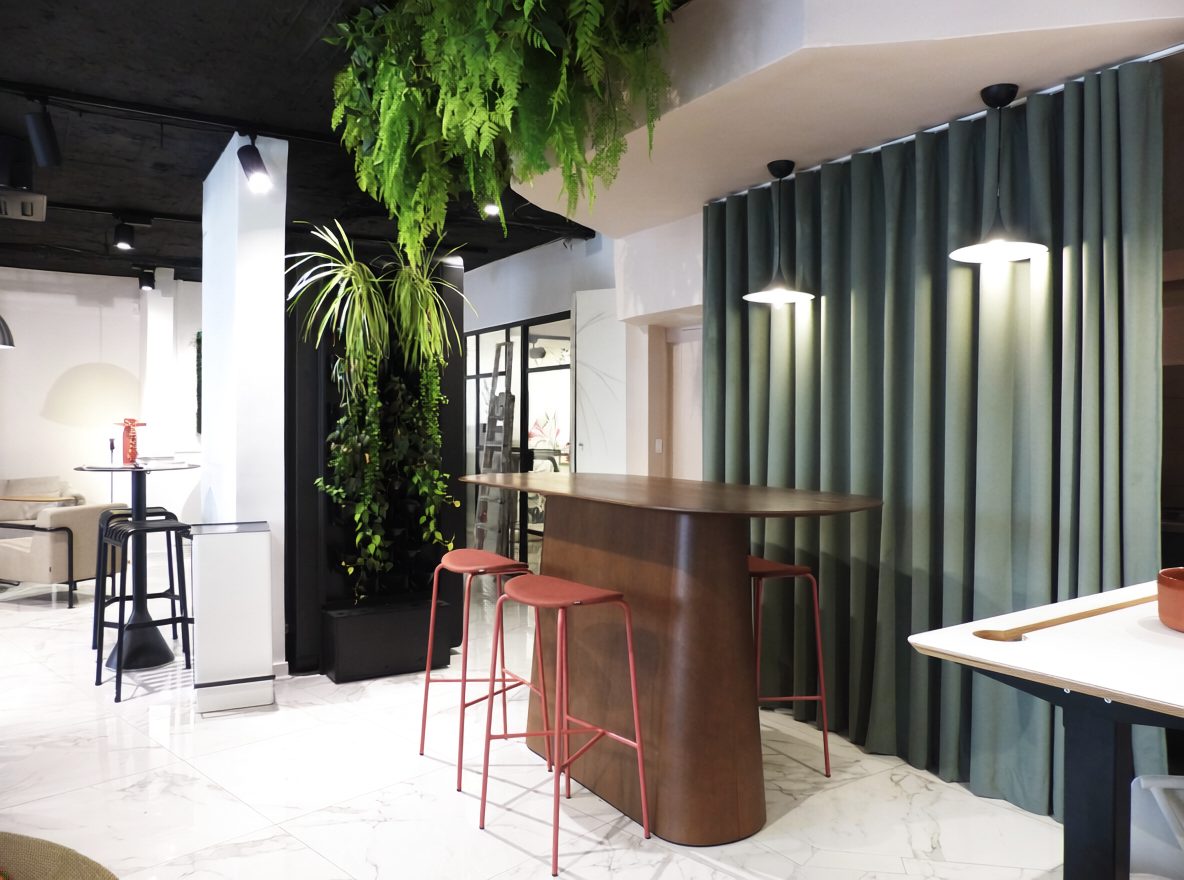 Accueil du showroom : comptoir haut arrondi, tabourets rouges et plantes suspendues